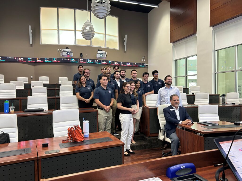 A group of students and a professor, dressed in business casual attire, pose for a photo in a modern classroom with white chairs, wood desks, and digital ticker displays on the walls.