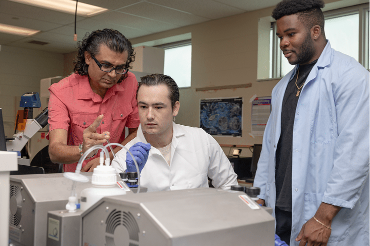 Three men in lab coats and gloves work together in a laboratory, with one man pointing and explaining equipment to the others who are observing attentively.