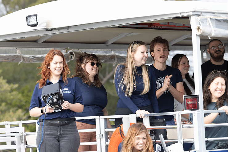 A group of smiling people stand together on the deck of a boat under a canopy, some wearing navy blue shirts. One woman holds a camera or remote device. Trees are visible in the background.