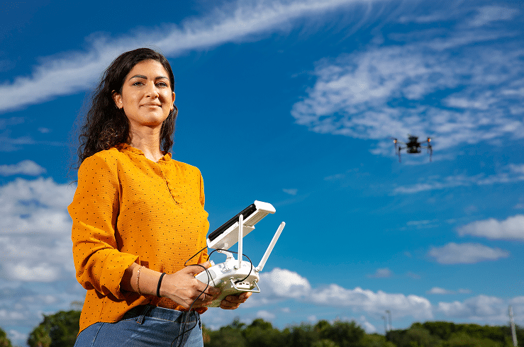 A woman in a yellow blouse stands outdoors holding a drone controller, with a flying drone visible in the sky against a backdrop of blue sky and scattered clouds.