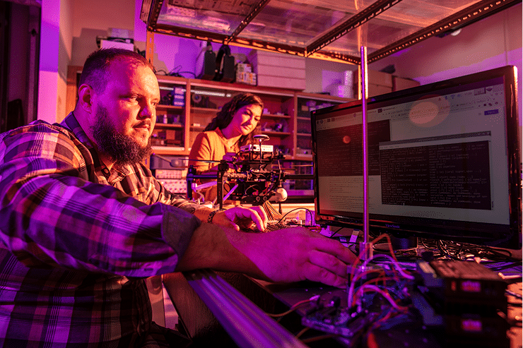 Two people work in a dimly lit room with purple lighting, surrounded by computers, wires, and electronic equipment. One is typing on a keyboard while the other smiles at their own workstation in the background.
